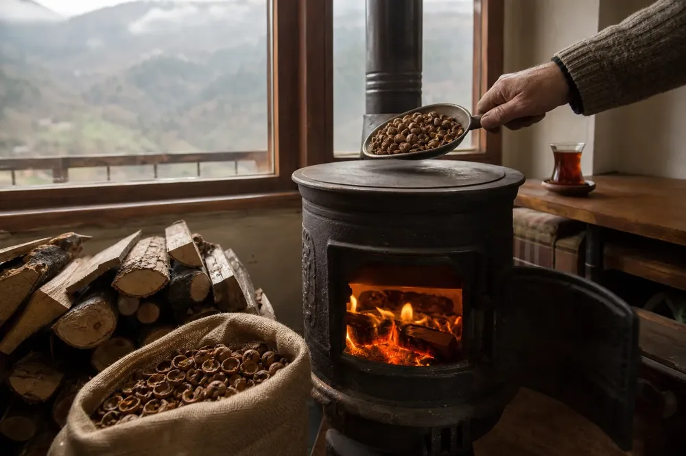 A hand adds hazelnut shells to a burning stove in a Black Sea home, with a shell sack and tea glass